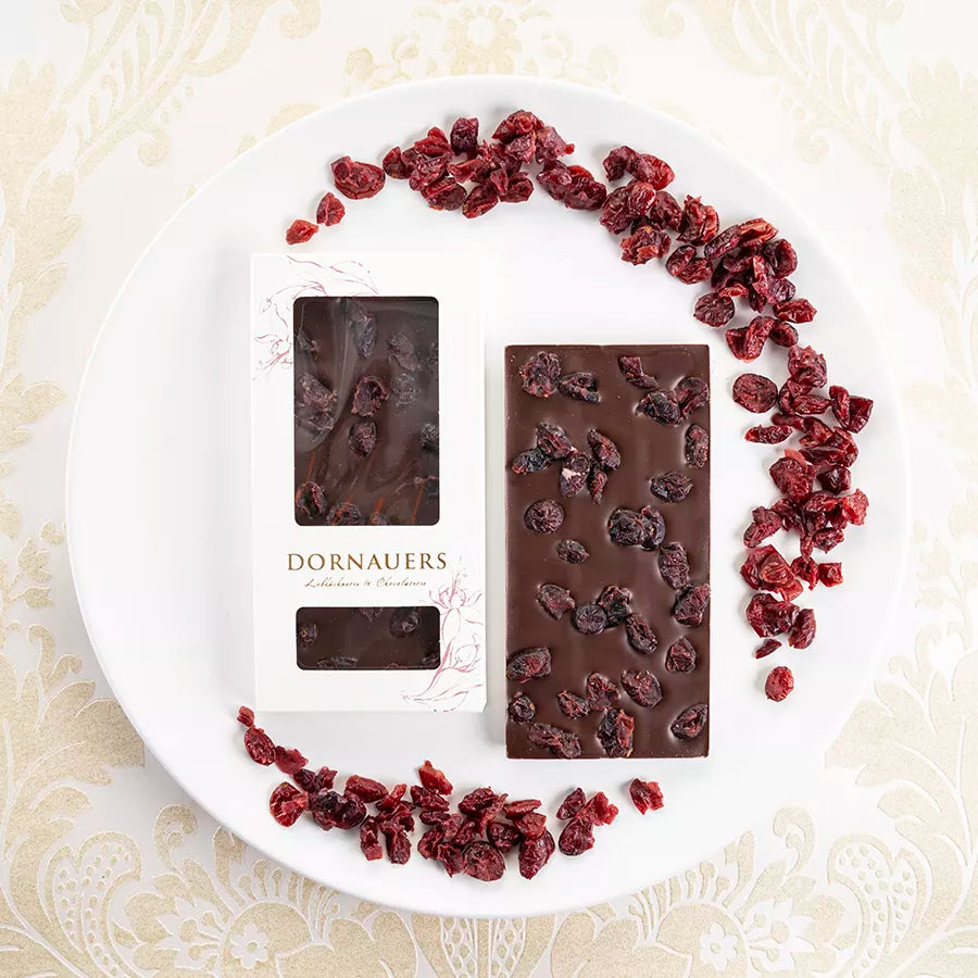 Dornauers Grand Cru Schokolade Cranberry dunkel auf weißem Teller, daneben die Verpackung und getrocknete Cranberrystücke