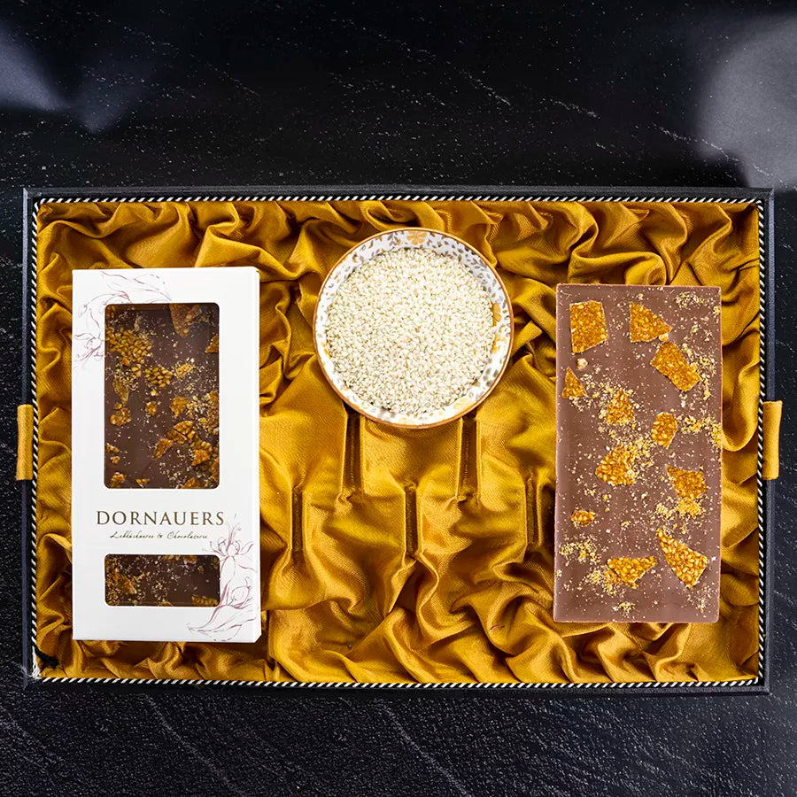 DORNAUERS Grand Cru Schokolade Vollmilch Sesam Karamell daneben eine Schale voll Sesam und die Verpackung auf einem goldenem Tuch