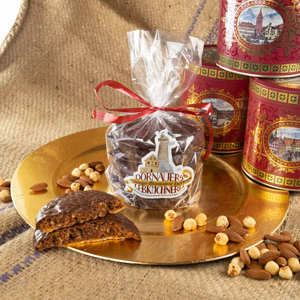 Dornauers 5er Pack Elisen-Lebkuchen dunkle Schokoladenkuvertüre in transparenter Folie mit roter Schleife auf einem goldenen Teller, umgeben von Haselnüssen, Mandeln und roten Blechdosen mit historischen Stadtmotiven