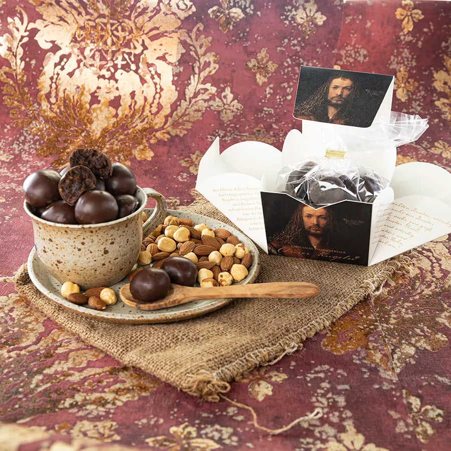 Dornauers Original Nürnberger Dürer Kugeln in einer geöffneten Verpackung mit Dürrer-Porträt, daneben eine Tasse Voller Dürrer Kugeln, Haselnüsse, Mandeln und ein Holzlöffel auf einem Keramikteller