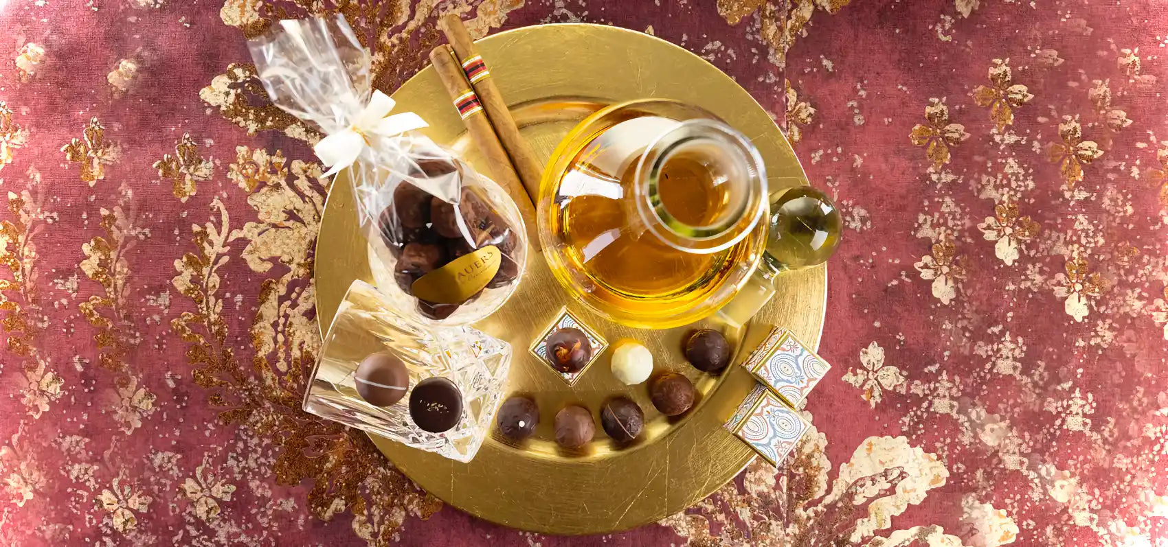 Arrangement aus Dornauers edlen Pralinen, Zigarren, einem umgekippten Glas, transparenter Geschenkverpackung und einer Glaskanne mit Rum auf goldenem Teller.