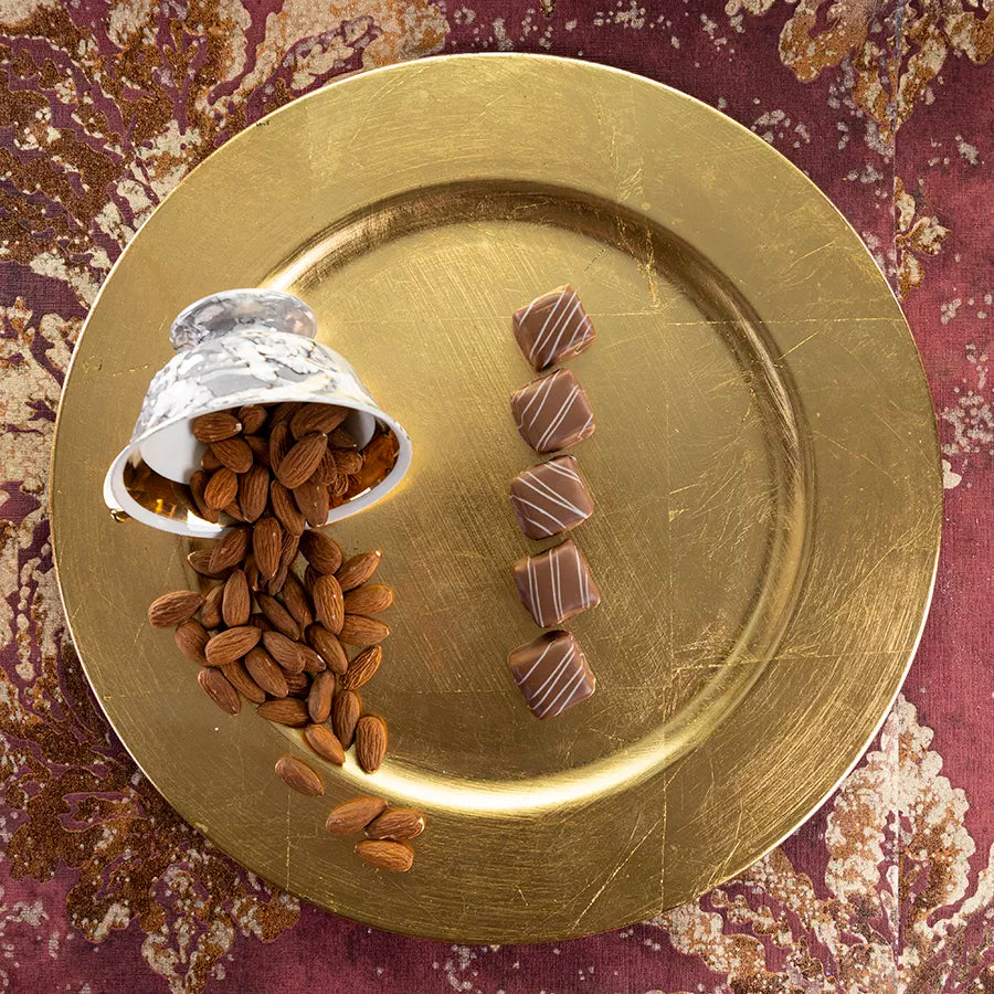 DORNAUERS Grand Cru Pralinen Marzipan Karamell auf goldenem Teller, daneben eine umgekippte Tasse mit ganzen Mandeln 