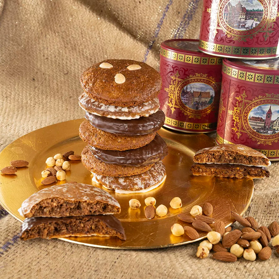 Turm aus verschiedenen Elisenlebkuchen – naturbelassen, schokoladenüberzogen und süß glasiert – auf goldenem Teller, umgeben von Haselnüssen, Mandeln und roten Blechdosen mit historischen Stadtmotiven