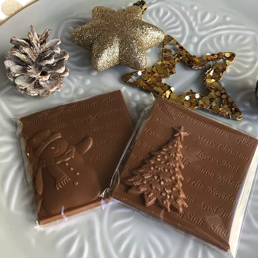 Dornauers Weihnachtstafeln Vollmilch mit Schneemann- und Tannenbaum-Motiv auf einem weißen Teller, dekoriert mit Zapfen und goldenen Sternen