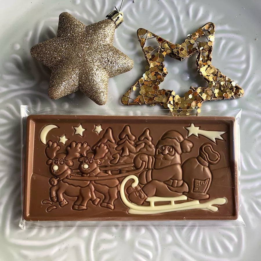 Dornauers Weihnachtstafel Vollmilch mit Weihnachtsmann im Schlitten und Rentieren Motiv, verziert mit weißer Schokolade, auf einem weißen Teller dekoriert mit goldenen Sternen  