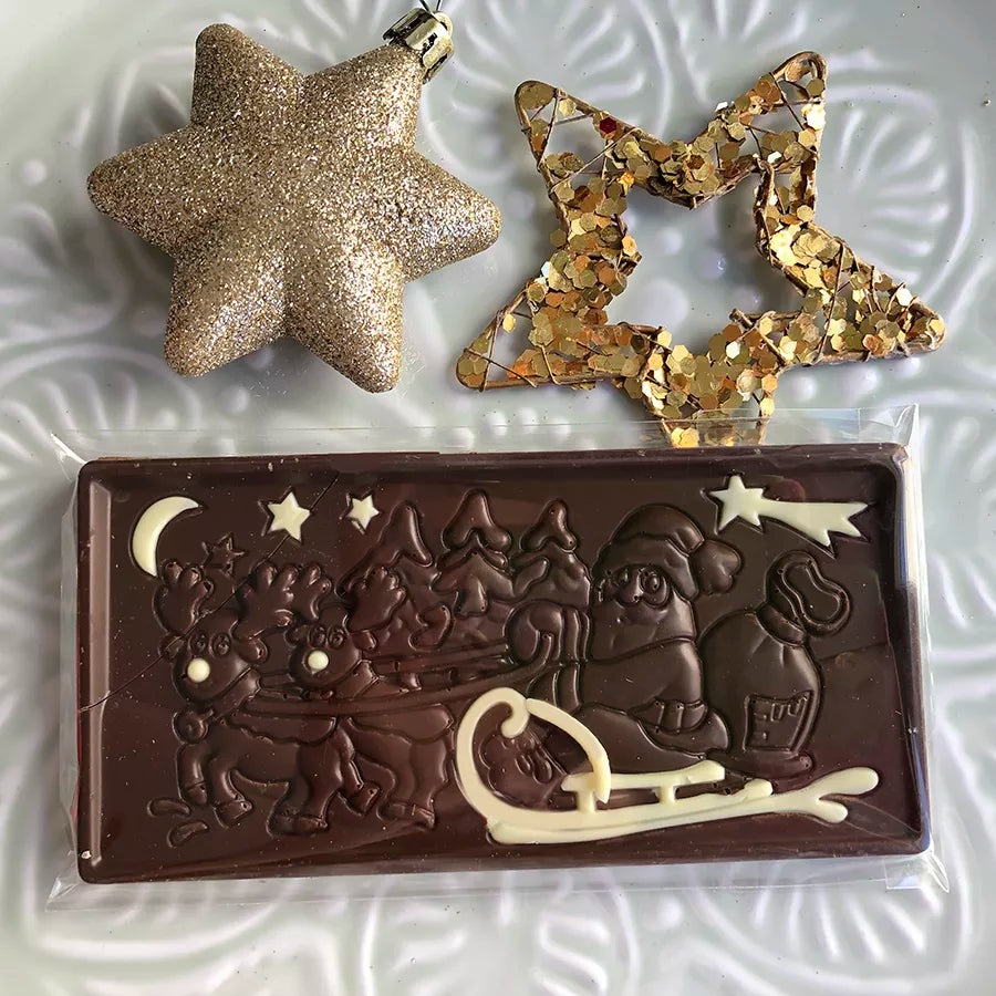 Dornauers Weihnachtstafel Zartbitter mit Weihnachtsmann im Schlitten und Rentieren Motiv, verziert mit weißer Schokolade, auf einem weißen Teller dekoriert mit goldenen Sternen  