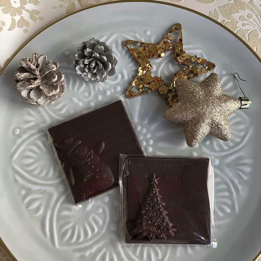 Dornauers Weihnachtstafeln Zartbitter mit Schneemann- und Tannenbaum-Motiv auf einem weißen Teller, dekoriert mit Zapfen und goldenen Sternen